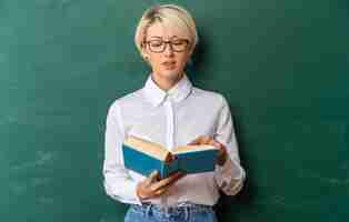 Free photo pleased young blonde female teacher wearing glasses in classroom standing in front of chalkboard holding pointing finger on and reading book with copy space