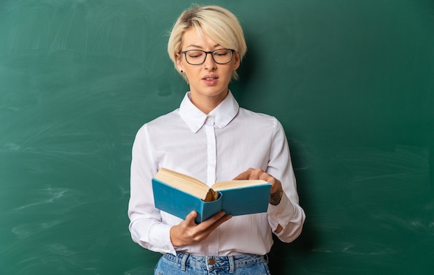 pleased young blonde female teacher wearing glasses in classroom standing in front of chalkboard holding pointing finger on and reading book with copy space