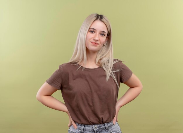 Free photo pleased young blonde girl putting hands on waist on isolated green space with copy space