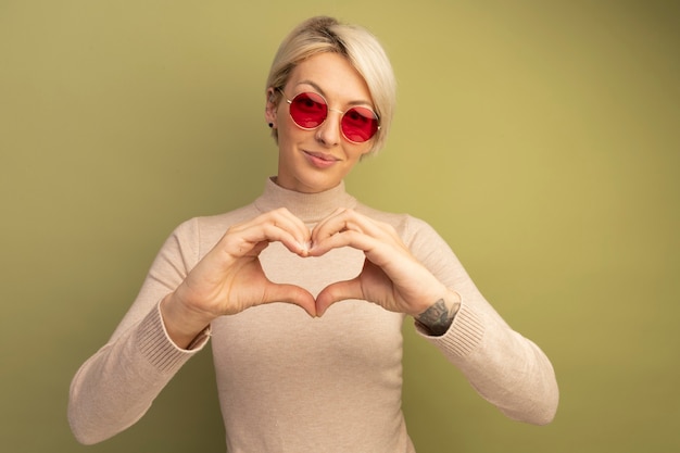 Pleased young blonde girl wearing sunglasses doing heart sign 