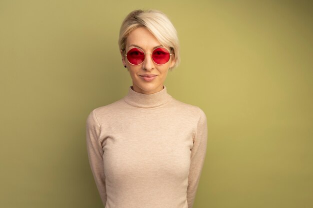 Pleased young blonde girl wearing sunglasses keeping hands behind back isolated on olive green wall with copy space