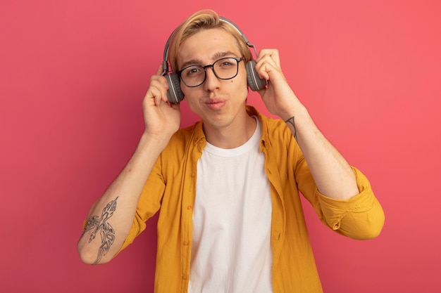 Pleased young blonde guy looking straight ahead wearing yellow t-shirt and glasses with headphones isolated on pink