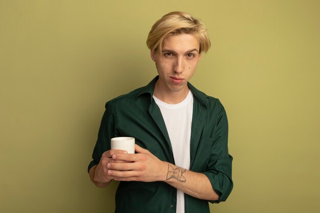 Довольный молодой блондин в зеленой футболке, скрестив руки с чашкой чая