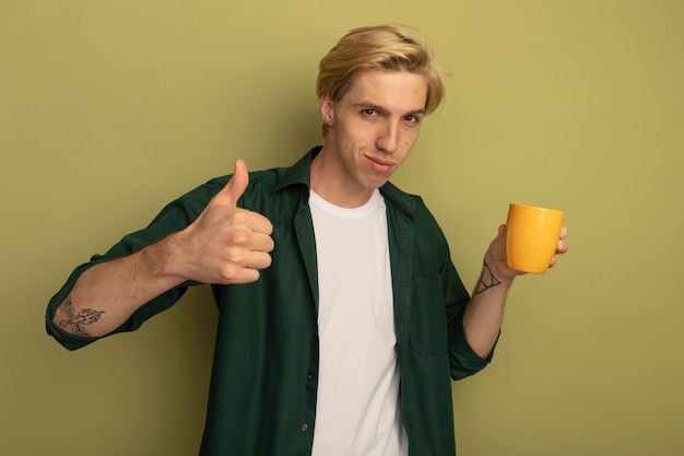 Soddisfatto giovane ragazzo biondo che indossa la maglietta verde che tiene la tazza di tè e che mostra il pollice in su