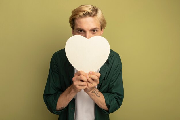 Pleased young blonde guy wearing green t-shirt holding heart shape box