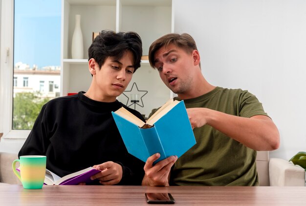Pleased young blonde handsome man holds and points at book sitting at table with young brunette handsome boy looking at book inside living room