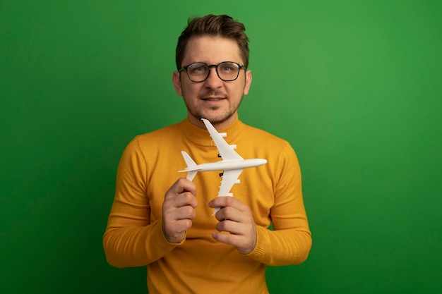 Pleased young blonde handsome man wearing glasses holding model plane isolated on green wall with copy space