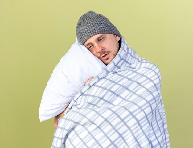 Pleased young blonde ill man wearing winter hat and scarf wrapped in plaid hugs pillow isolated on olive green wall