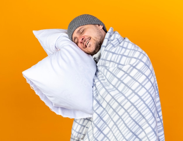 Pleased young blonde ill man wearing winter hat wrapped in plaid holds and puts head on pillow isolated on orange wall