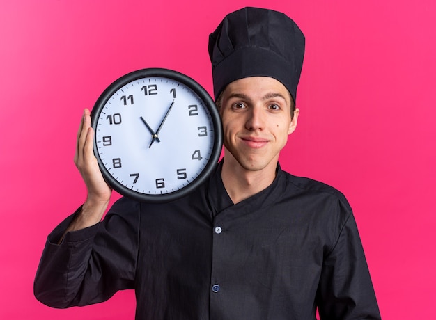 Pleased young blonde male cook in chef uniform and cap holding clock touching face with it looking at camera isolated on pink wall