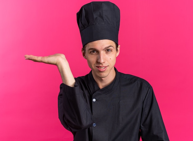 Pleased young blonde male cook in chef uniform and cap showing empty hand 