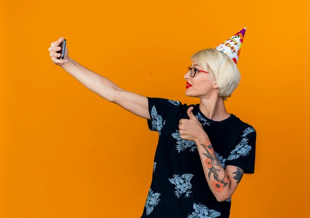 Довольная молодая блондинка тусовщица в очках и кепке на день рождения, делающая селфи, показывая большой палец вверх изолированно на оранжевом фоне