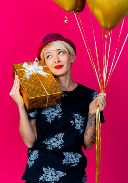 Pleased young blonde party girl wearing party hat holding gift box on shoulder and balloons looking at side isolated on crimson background