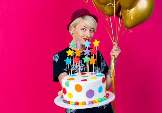 Pleased young blonde party girl wearing party hat looking at camera holding balloons and stretching out birthday cake with stars towards camera isolated on crimson background with copy space