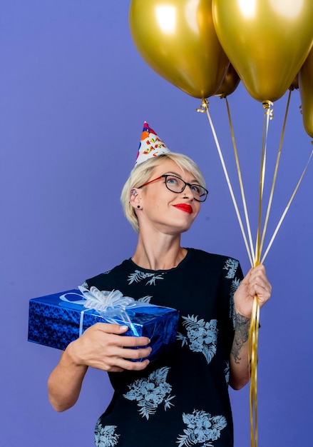 Pleased young blonde party woman wearing glasses and birthday cap holding balloons and gift box looking up plunging into dreams isolated on purple wall