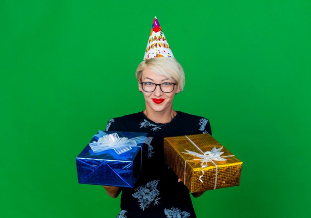 Pleased young blonde party woman wearing glasses and birthday cap holding gift boxes looking at front isolated on green wall with copy space