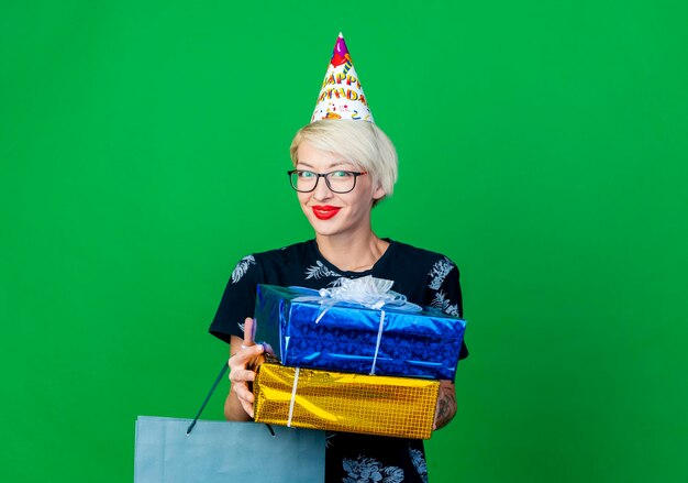 コピースペースで緑の壁に隔離された正面を見て、紙袋とギフトボックスを保持している眼鏡と誕生日キャップを身に着けている若い金髪のパーティーの女性を喜ば