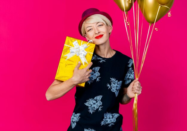 Felice giovane bionda festa donna che indossa il cappello del partito che tiene il contenitore di regalo e palloncini guardando la parte anteriore isolata sulla parete rosa con lo spazio della copia