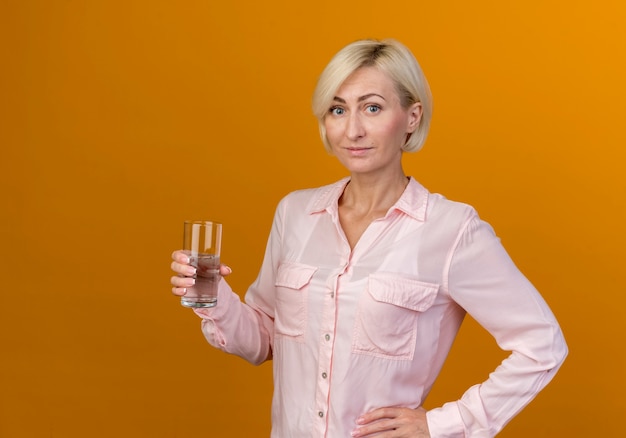 Free photo pleased young blonde slavic woman holding glass of water and putting hand on hip