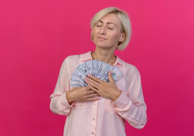 Pleased young blonde slavic woman holding money with closed eyes isolated on pink background with copy space