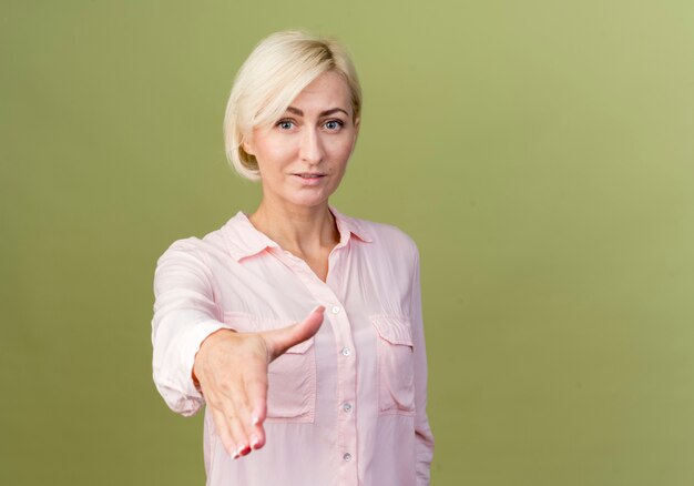 Pleased young blonde slavic woman holding out hand  isolated on olive green wall