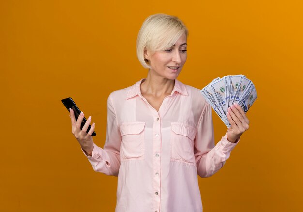 Pleased young blonde slavic woman holding phone and looking at cash in her hand 