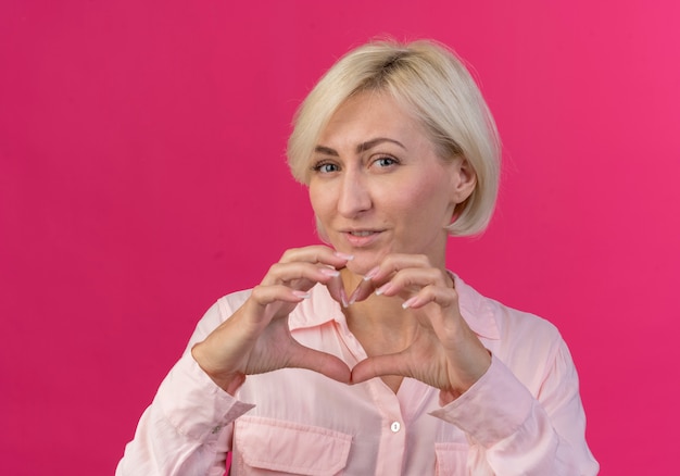 Довольная молодая блондинка славянская женщина смотрит в камеру и делает знак сердца, изолированные на розовом фоне