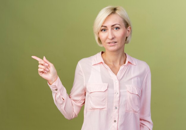 Pleased young blonde slavic woman points at side isolated on olive green wall with copy space