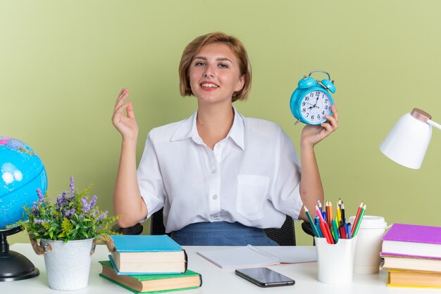 Pleased young blonde student girl sitting at desk with school tools keeping hand in air holding alarm clock looking at camera isolated on olive green wall