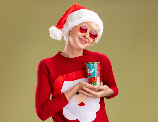 Pleased young blonde woman wearing christmas hat and santa claus christmas sweater with glasses holding plastic christmas cup with closed eyes isolated on olive green background with copy space