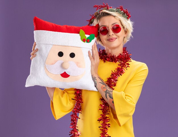 보라색 벽에 고립 된 닫힌 된 눈으로 산타 클로스 베개를 들고 목에 크리스마스 머리 화 환과 반짝이 갈 랜드를 입고 기쁘게 젊은 금발의 여자