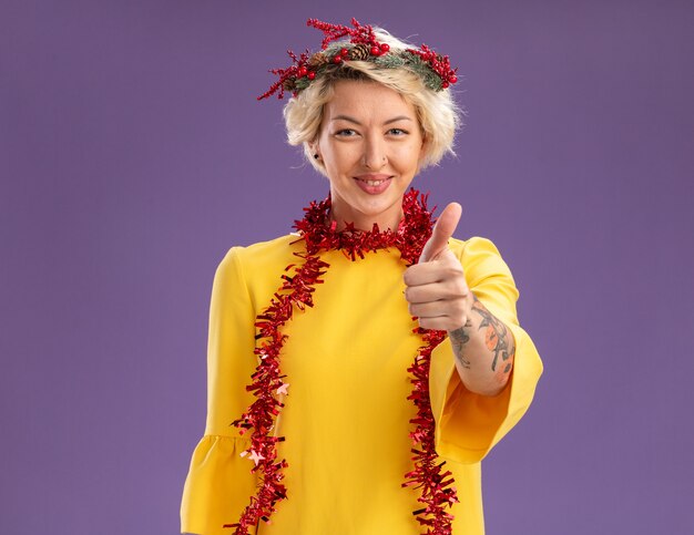 Free photo pleased young blonde woman wearing christmas head wreath and tinsel garland around neck looking  showing thumb up isolated on purple wall with copy space