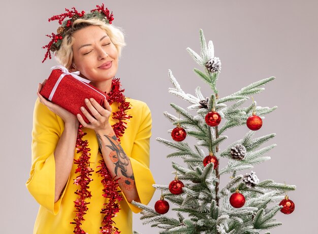 クリスマス ヘッド リースと見掛け倒しの花輪を身に着けている幸せな若いブロンドの女性が飾られたクリスマス ツリーの近くに立って、白い壁に目を閉じてギフト パッケージを保持しています。