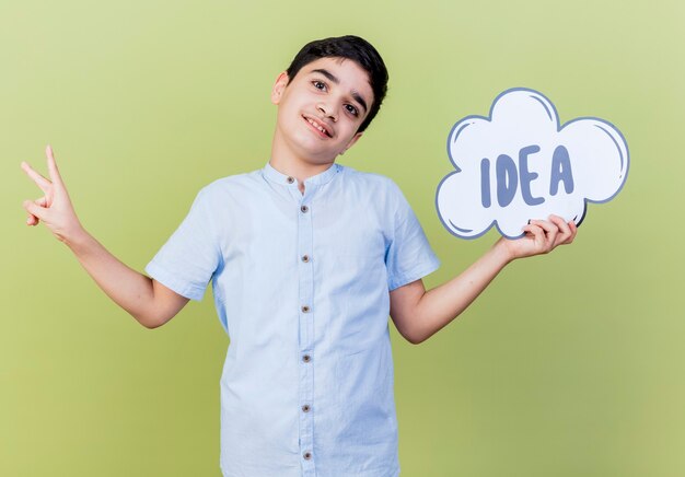 Pleased young boy holding idea bubble looking at front doing peace sign isolated on olive green wall