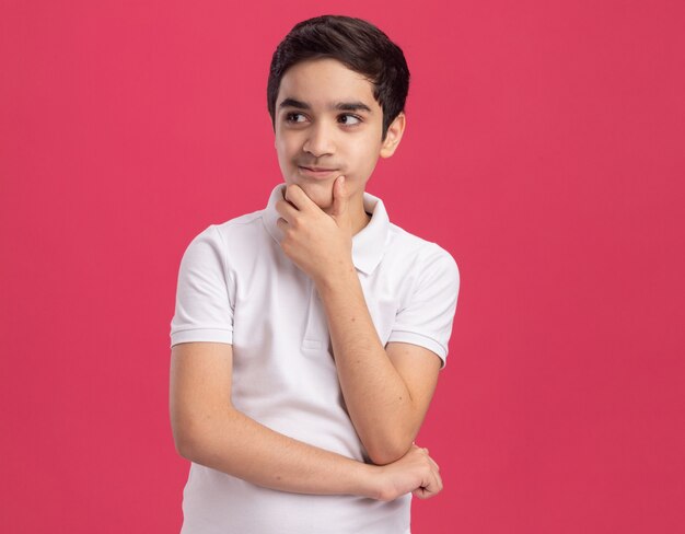 Pleased young boy touching chin looking at side isolated on pink wall with copy space