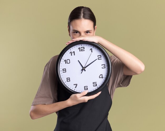 Pleased young brunette barber girl in uniform holds clock on olive green