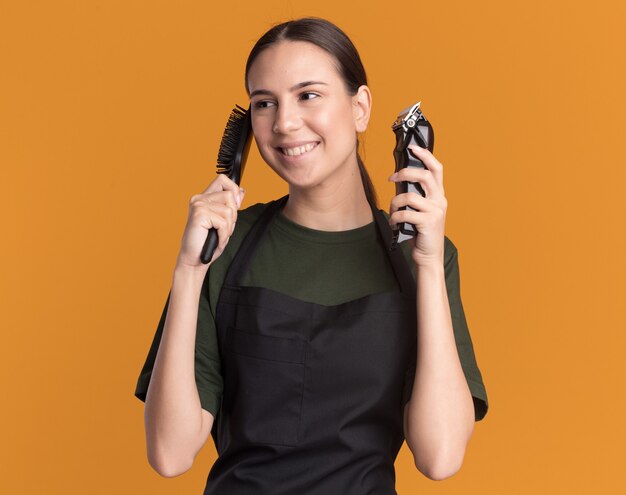 Pleased young brunette barber girl in uniform holds hair clippers and comb looking at side on orange