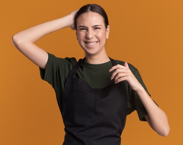 Pleased young brunette barber girl in uniform puts hand on head behind and looks at camera on orange