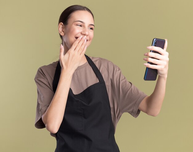 Pleased young brunette barber girl in uniform puts hand on mouth holding and looking at phone isolated on olive green wall with copy space