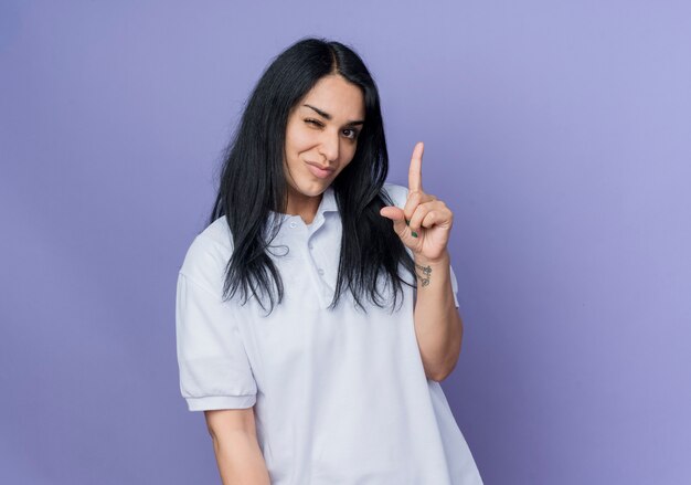 Pleased young brunette caucasian girl blinks eye and points up isolated on purple wall