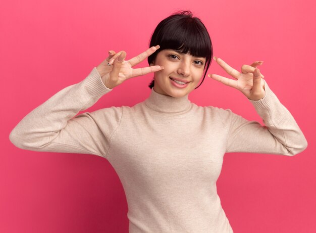 Pleased young brunette caucasian girl gestures victory sign with two hands on pink