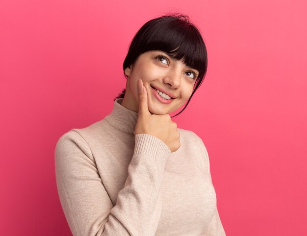 Pleased young brunette caucasian girl holds chin and looks up