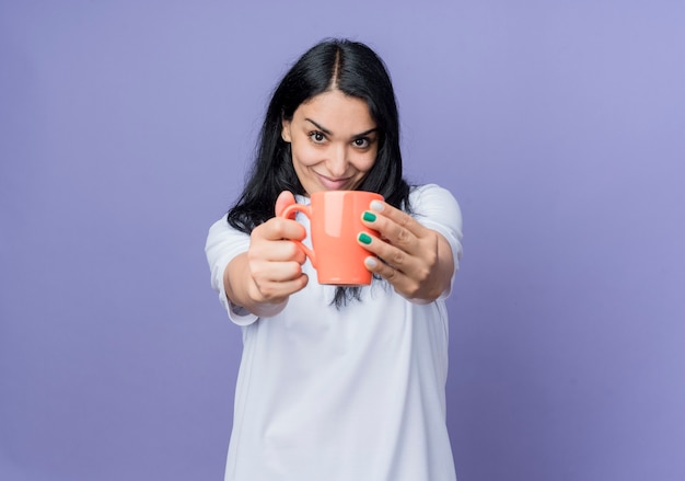 Pleased young brunette caucasian girl holds out cup isolated on purple wall