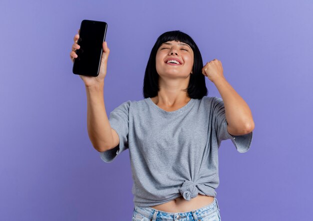 Pleased young brunette caucasian girl holds phone and raises fist 