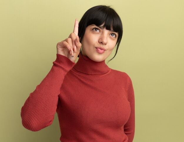 Pleased young brunette caucasian girl points and looks up isolated on olive green wall with copy space
