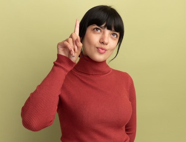 Pleased young brunette caucasian girl points and looks up isolated on olive green wall with copy space