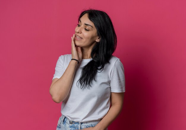 Pleased young brunette caucasian girl puts hand on mouth looking at side isolated on pink wall