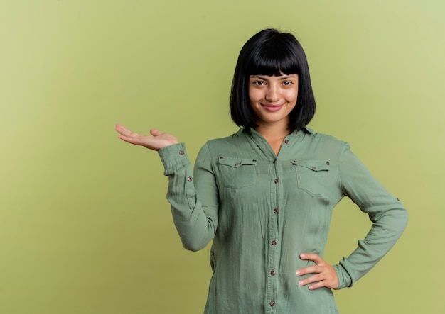 Pleased young brunette caucasian girl puts hand on waist and holds hand open looking at camera isolated on olive green background with copy space