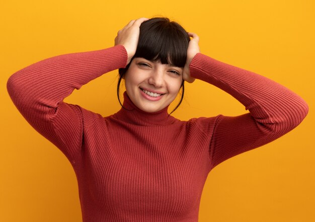 Pleased young brunette caucasian girl puts hands on head isolated on orange wall with copy space