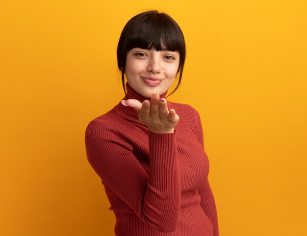 Pleased young brunette caucasian girl sends kiss with hand isolated on orange wall with copy space